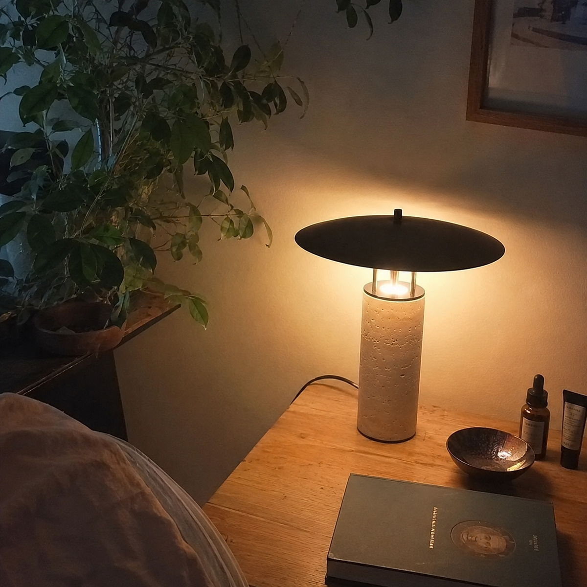 Lampe de chevet allumée photographiée par un client dans sa chambre, avec une table de nuit en bois et des objets personnels visibles dans une lumière tamisée. Image